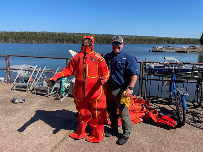 Two people stand on a cement dock overlooking a small harbor. The woman is wearing a full-body, orange neoprene suit with yellow seams. It is the kind of float-suit worn by people when abandoning a ship. The man next to her wears regular clothes.