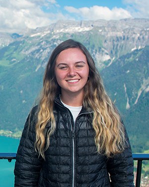 A woman with long blonde hair wearing a black puffy jacket stands in front of a railing with green/gray/white mountains behind her.