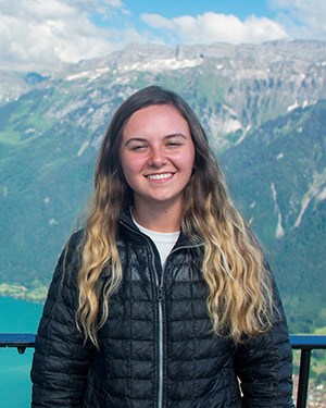 KateRoberts A woman with long blonde hair wearing a black puffy jacket stands in front of a railing with green/gray/white mountains behind her.