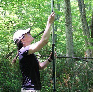 A woman wearing a baseball cap and purple t-shirt checks a microphone stand in the woods.