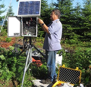 A man in a blue polo shirt stands in a forest opening, working on an electrical box below a solar panel.
