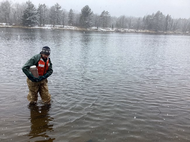 A person in a green coat and hat and wearing an orange life jacket walks through knee-high water carrying a clear jug of brownish water. Snowflakes fall around them, and the distant shore is covered in snow.