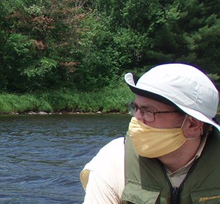 A man wearing a yellow face mask and a white hat looks the left. Water and trees are behind him.
