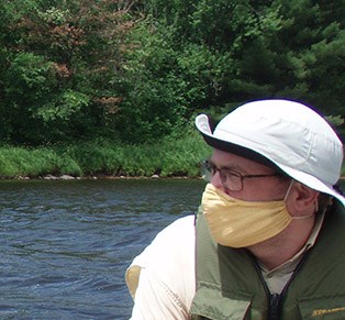 Thomas Parr A man wearing a yellow face mask and a white hat looks the left. Water and trees are behind him.