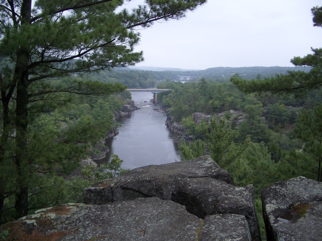 A distant highway bridge passes between two unbroken forests, high over a wide river. Grayish boulders and a single white pine tree are in the foreground.