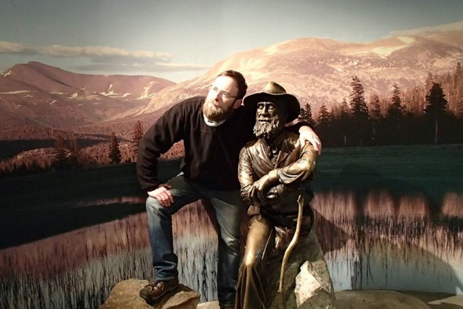 a man stands with his arm on the shoulders of a bronze statute