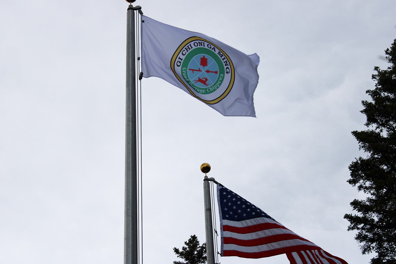 Photo of two flags, the U.S. flag on the right and the Grand Portage Ojibwe flag on the left. The Ojibwe flag is white with a round yellow ring around a green ring and a blue circle in the middle.