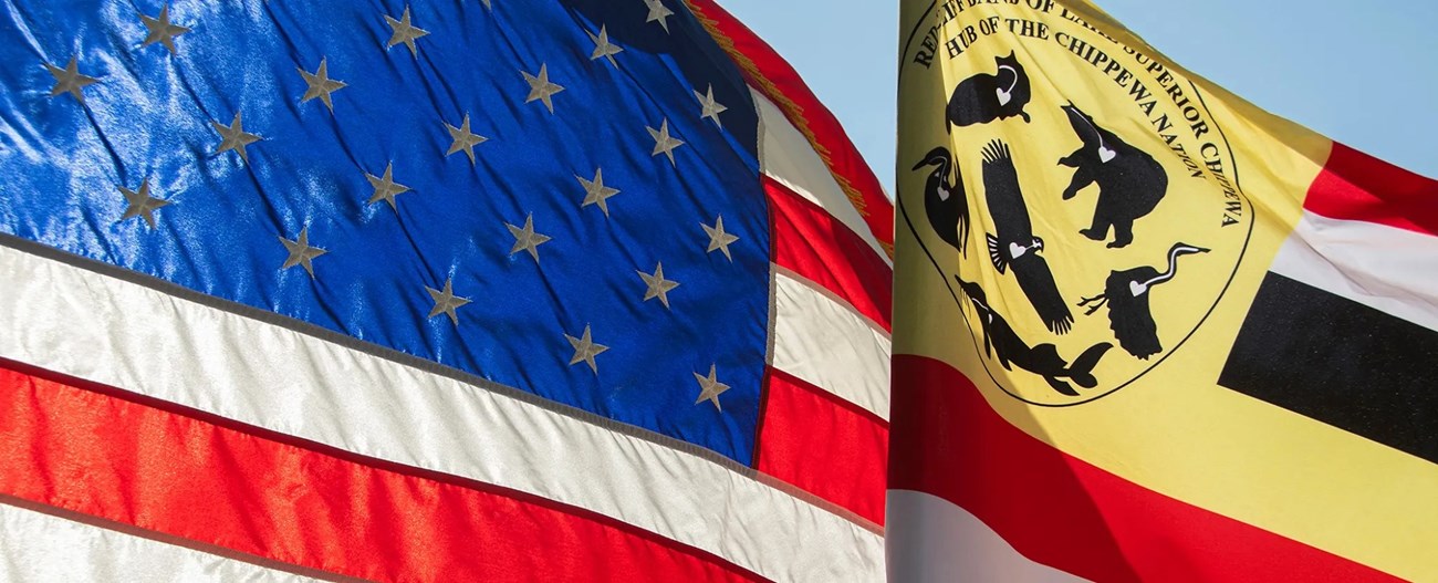 US-RedCliff Flags at APIS_cropped The red, white, and blue American flag flies next to a flag with a yellow field in the upper left corner and alternating red, white, black, and yellow horizontal stripes.