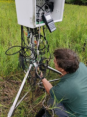 A man in a green polo shirt kneels in front of a mess of cords coming out of a metal box. The box is perched on a tripod with grass all around.