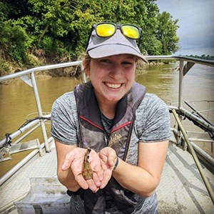 A scientist in a boat on a river holding a small fish in her hands smiling at the camera.