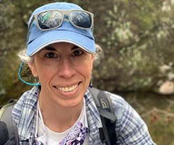 A close-up of a woman's head and shoulders. She is wearing a baseball cap and a backpack.