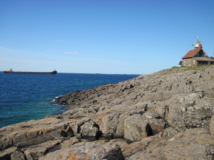 A long freighter floats near a rocky shoreline with a rock house.