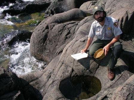 Bearded man in NPS uniform Crouches next to a rock pool holding a clipboard and round pan.