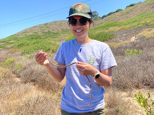 Linh Anh Cat, Ph.D. Person smiling on a sunny hillside, holding a snake.