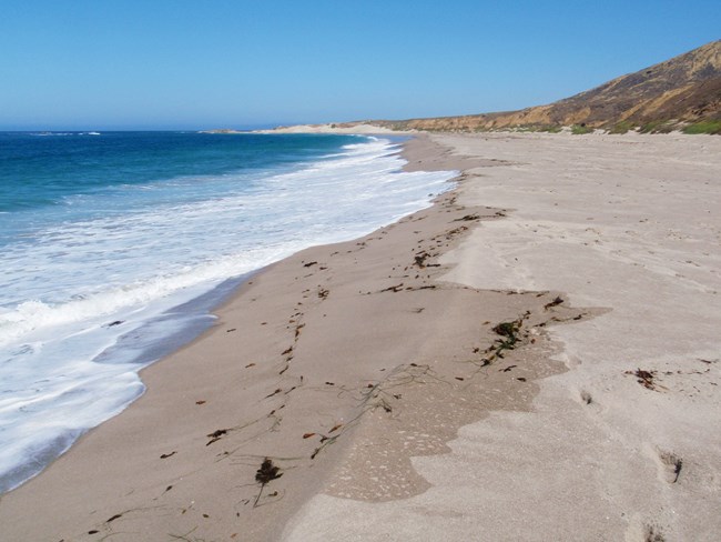 Small waves crash on a wide, sandy beach beneath a blue sky