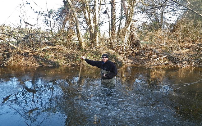Monitoring water quality in Wilderness Run, Fredericksburg and Spotsylvania County Battlefields National Military Park Person wearing waders, standing with a measuring pole in the middle of a river