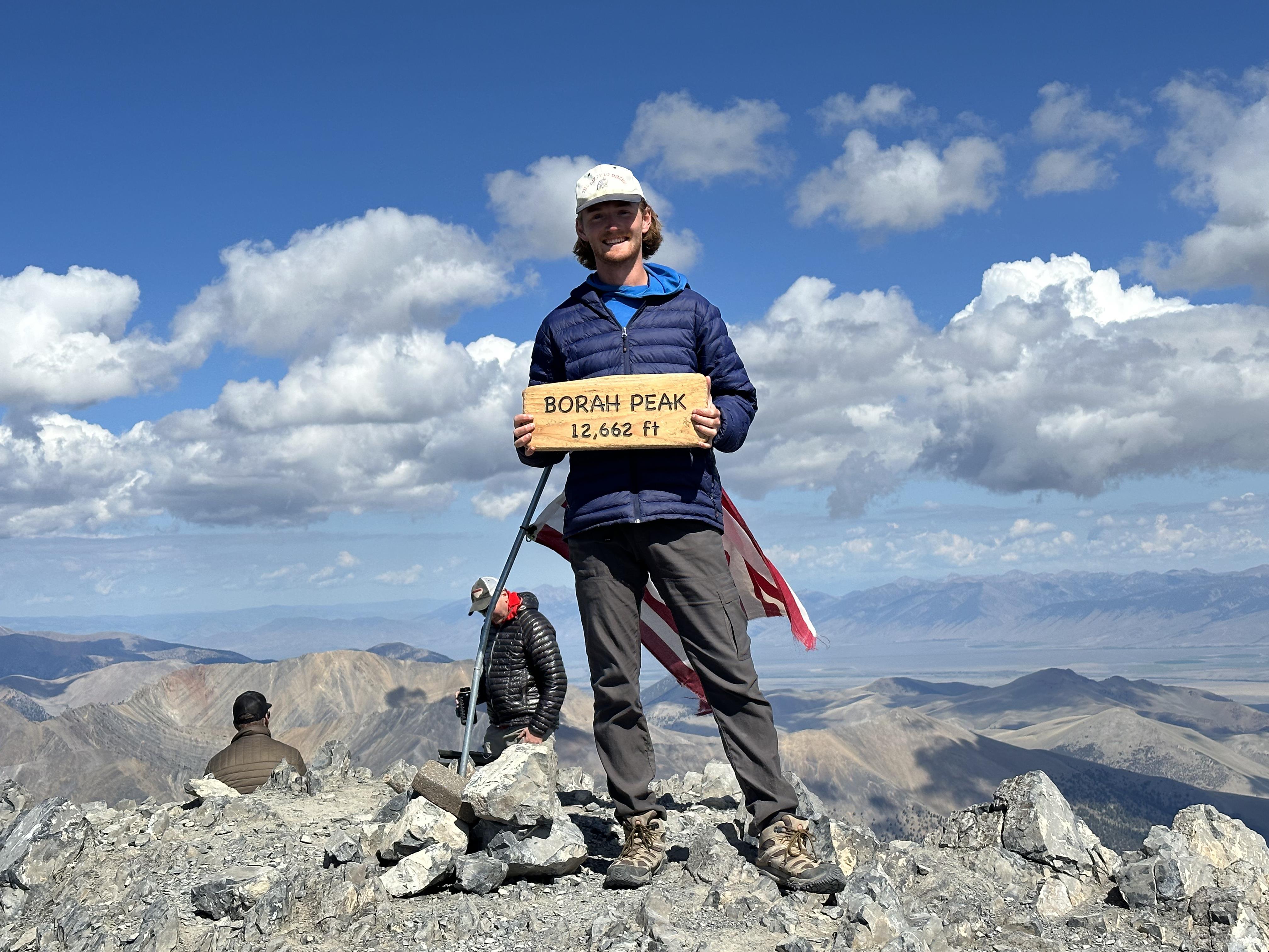 Man in puffy coat and baseball cap standing on rocky mountain summit and holding a sign that says "Borah Peak 12,662 ft."