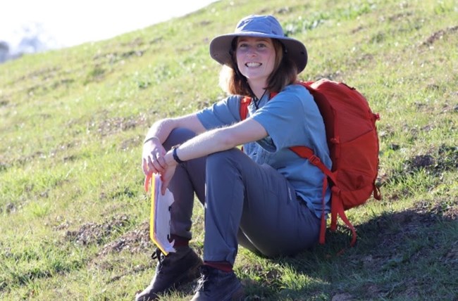 Woman wearing wide-brimed hat and daypack sitting on a sloped grassy area.