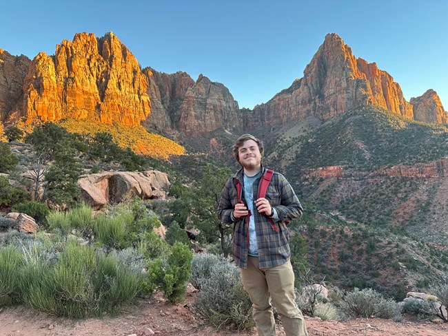 Smiling man wearing plaid shirt and daypack standing in arid landscape with morning-lit sandstone buttresses in background.