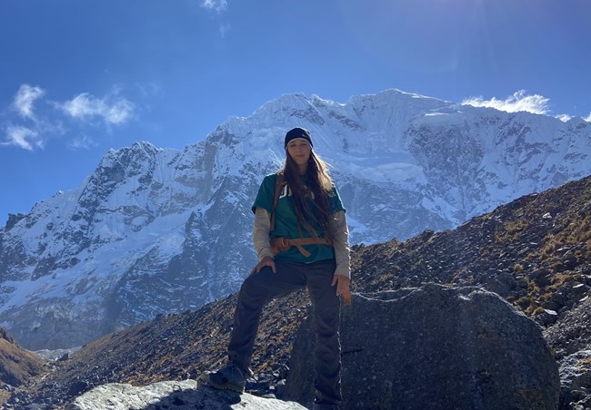 Ellie Bucci, Vegetation Technician Woman with long hair standing on a boulder with towering, snow covered mountain behind her.