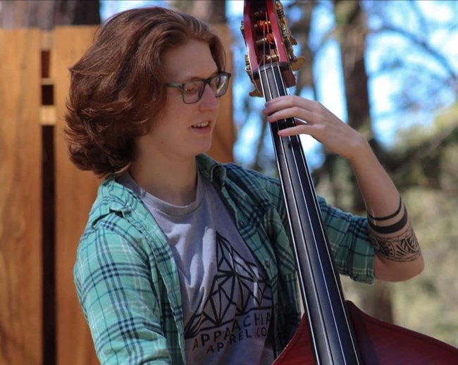 Em Michael, Physical Science Technician Person with wavy auburn hair and glasses holding a cello in outdoor setting.
