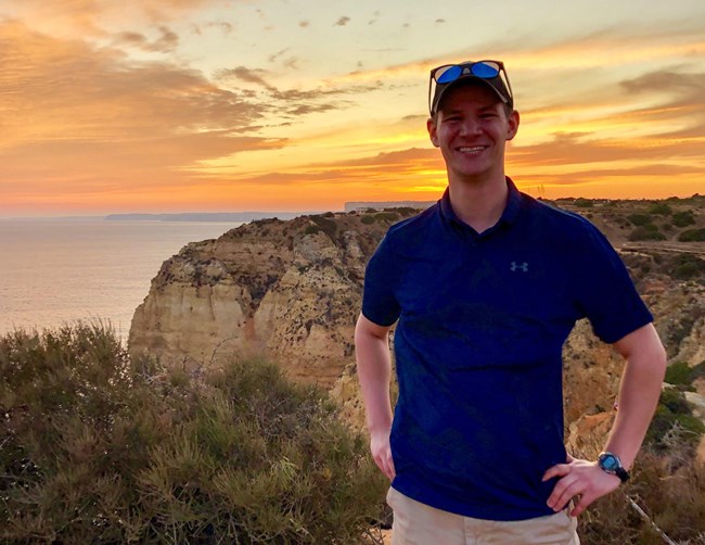 Man in blue shirt, hat, and sunglasses standing on bluff overlooking ocean at sunset