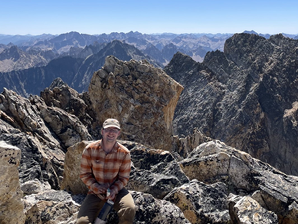 Man wearing long-sleeved shirt and ball cap sits amidst large rocks at the top of a peak with views of more peaks behind him.