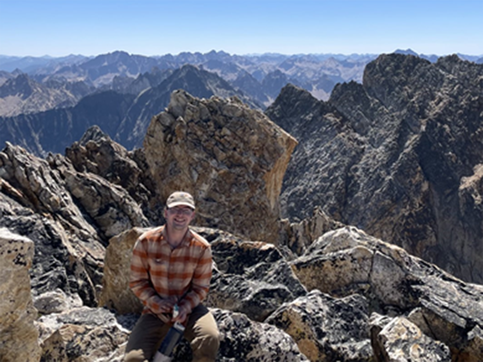 Gabe Elliot, Great Basin Institute Intern Man wearing long-sleeved shirt and ball cap sits amidst large rocks at the top of a peak with views of more peaks behind him.