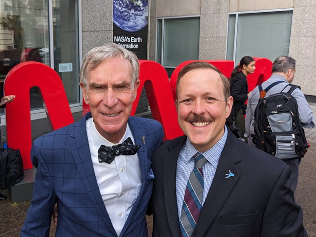 Two smiling men in blazers and neckties standing outside NASA headquarters.
