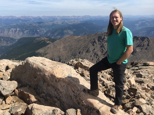 Kyle Smith, Biological Science Technician Man in green shirt standing on rocky summit with mountains in the distance