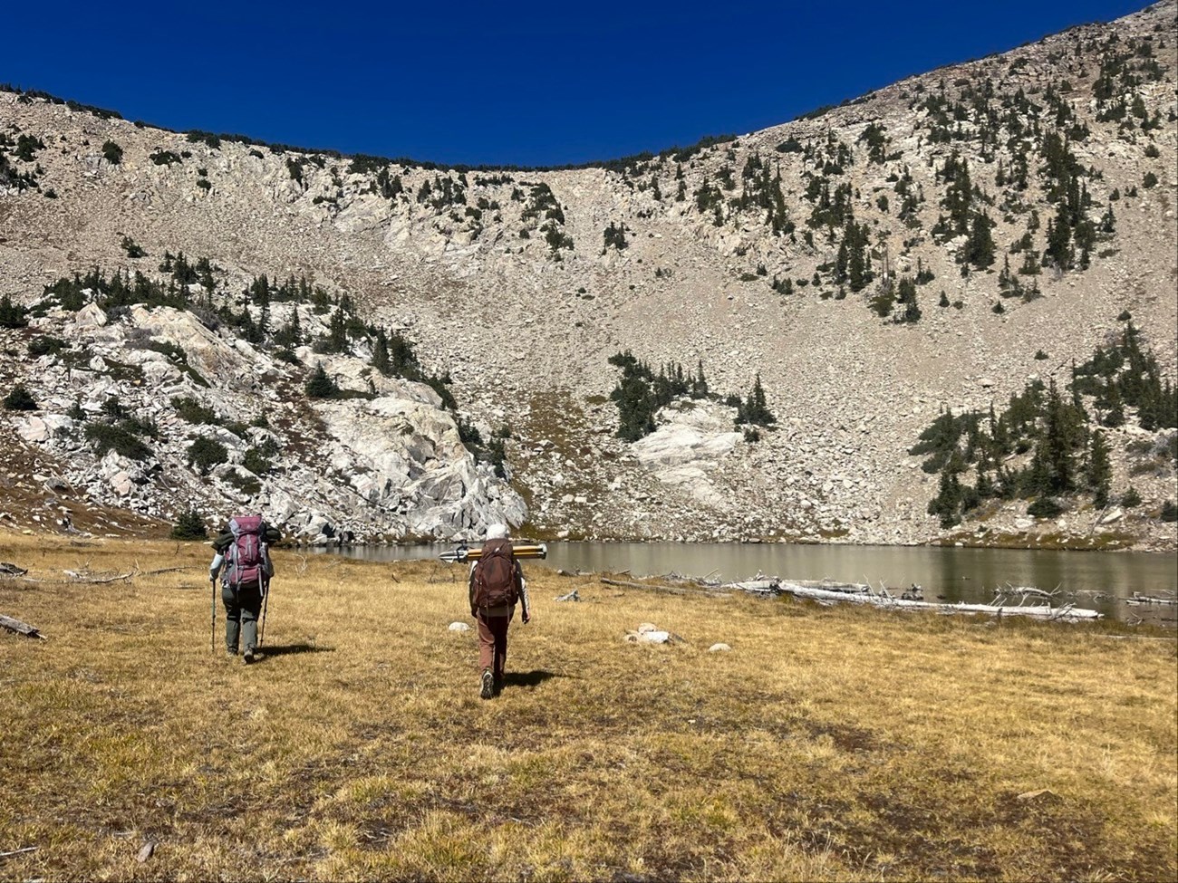 Two people with large backpacks and backs toward us hike towards a lake nestled at base of large talus slope, under cobalt blue sky.