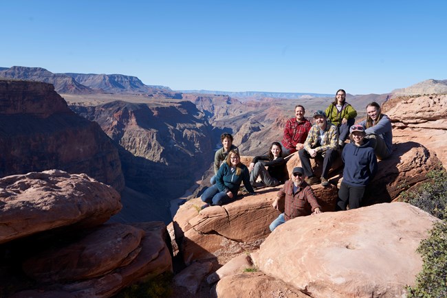 Nine people sitting in a group on red sandstone rock overlooking vast desert canyon.