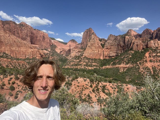 Man in white t-shirt with red sandstone rock formations and desert scrub vegetation, under azure sky, in the background.