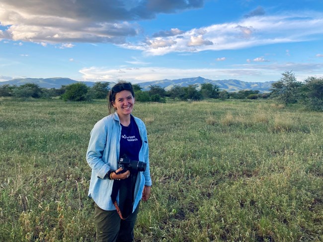 Smiling woman holding a large camera in an open field with mountains in the distance