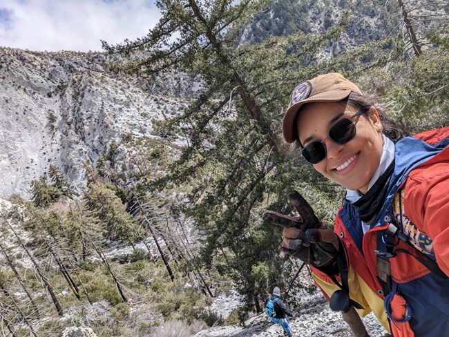 Smiling woman in a rocky, open-forested, mountain landscape.