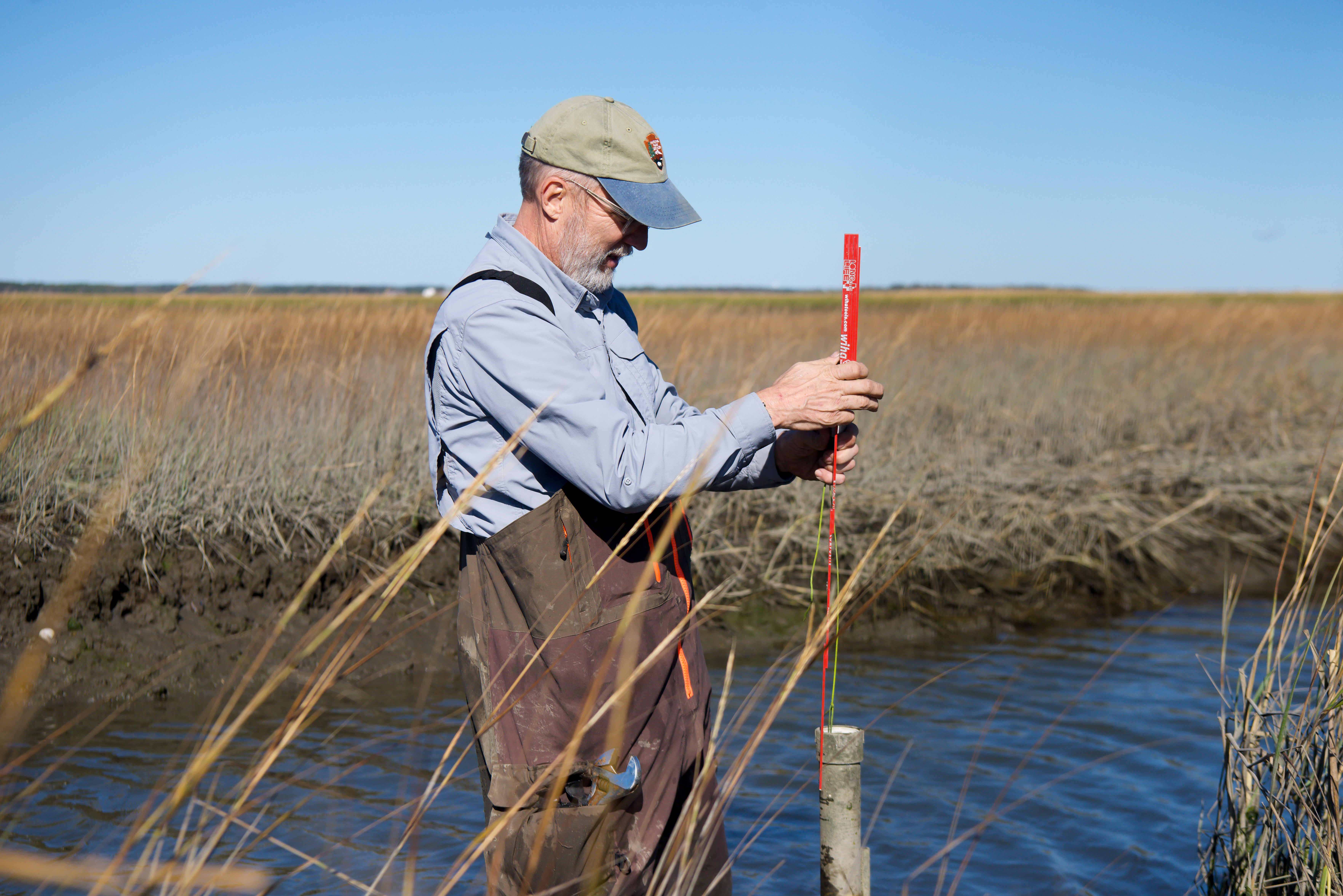 A field crew member stands holding a 6inch red rod over a pipe in a river