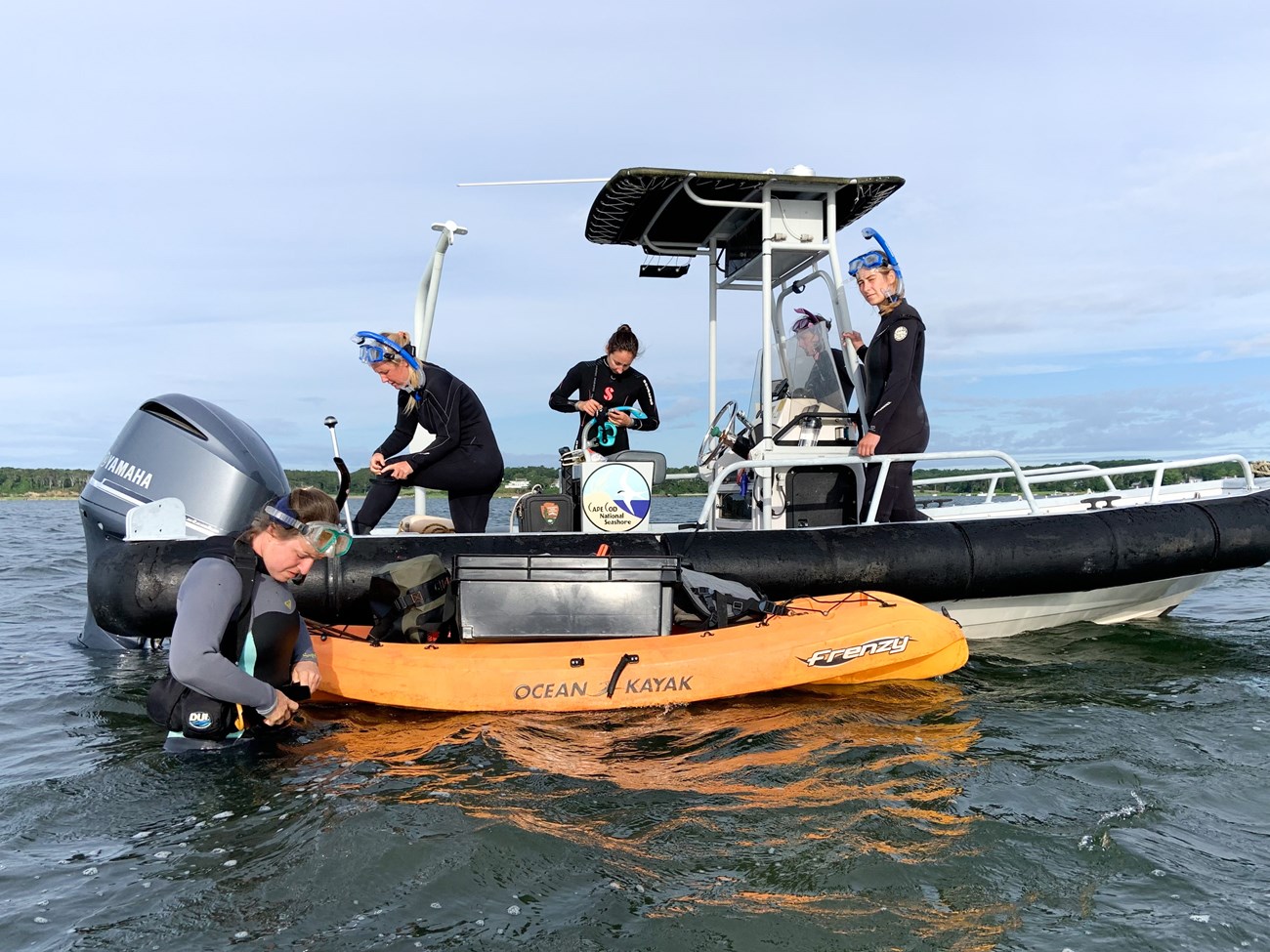 Four field crew members wearing wetsuits and snorkels stand on a boat. One researcher stands waist-deep, next to a yellow raft