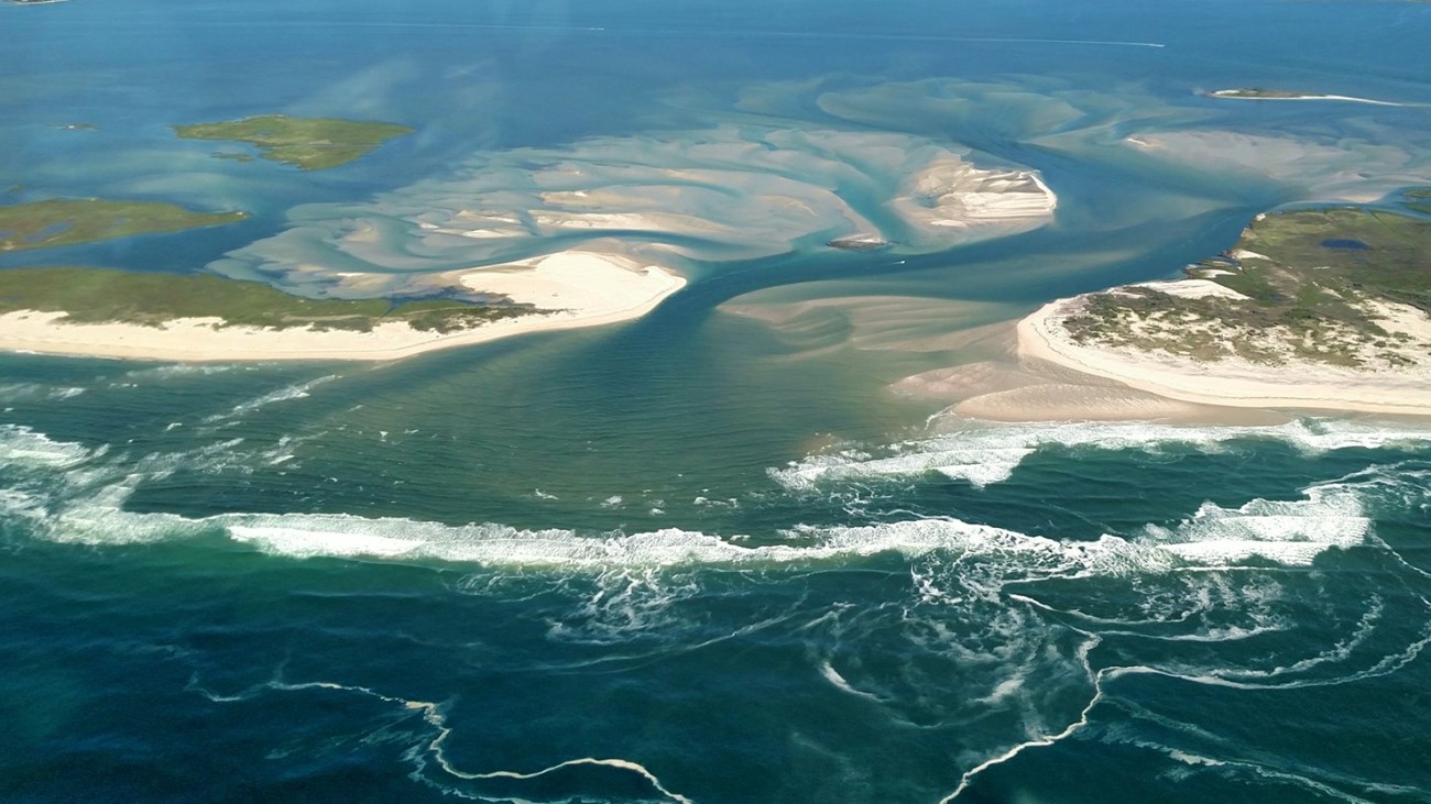 An aerial view of a long sandy island with a gap. Sand fans out from the gap in swirls.