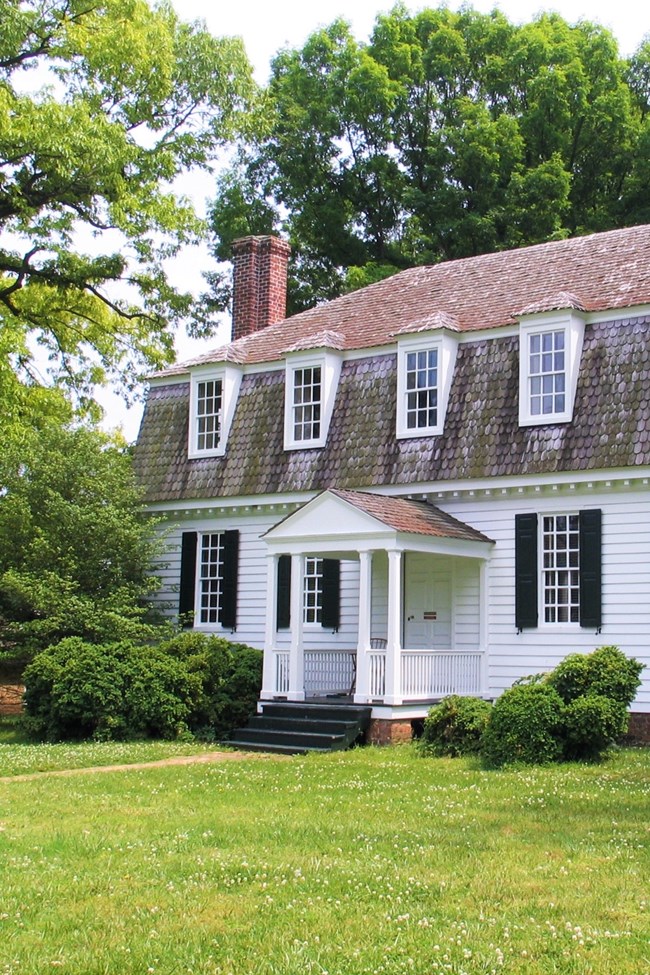 Moore House at COLO A two-story white stately house with brown barn roof and chimney stands on a lawn with trees