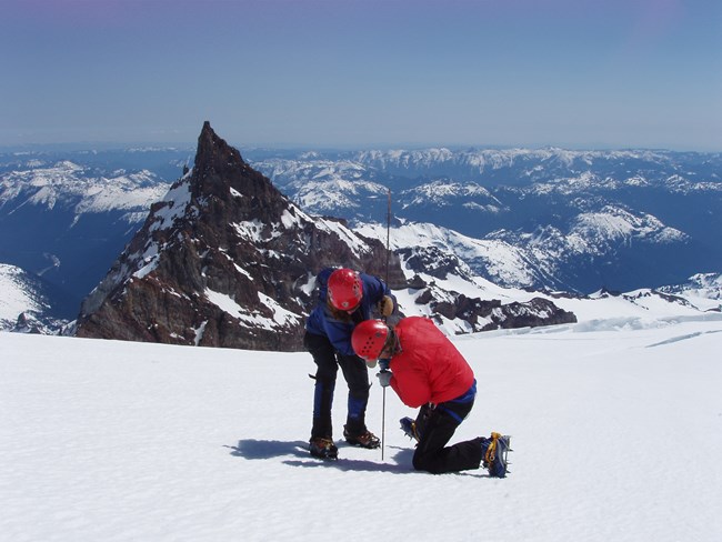 lt= Two people in winter clothing probing a glacier to measure snow depth with snowy peaks in background