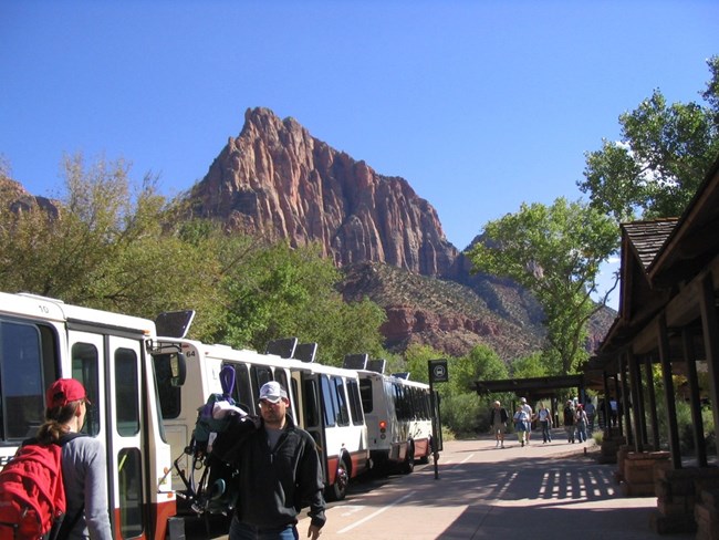 The shuttle system at Zion National Park helps manage high visitation and reduce vehicle impacts. Visitors board the park shuttle at Zion’s Visitor Center beneath towering red cliffs.
