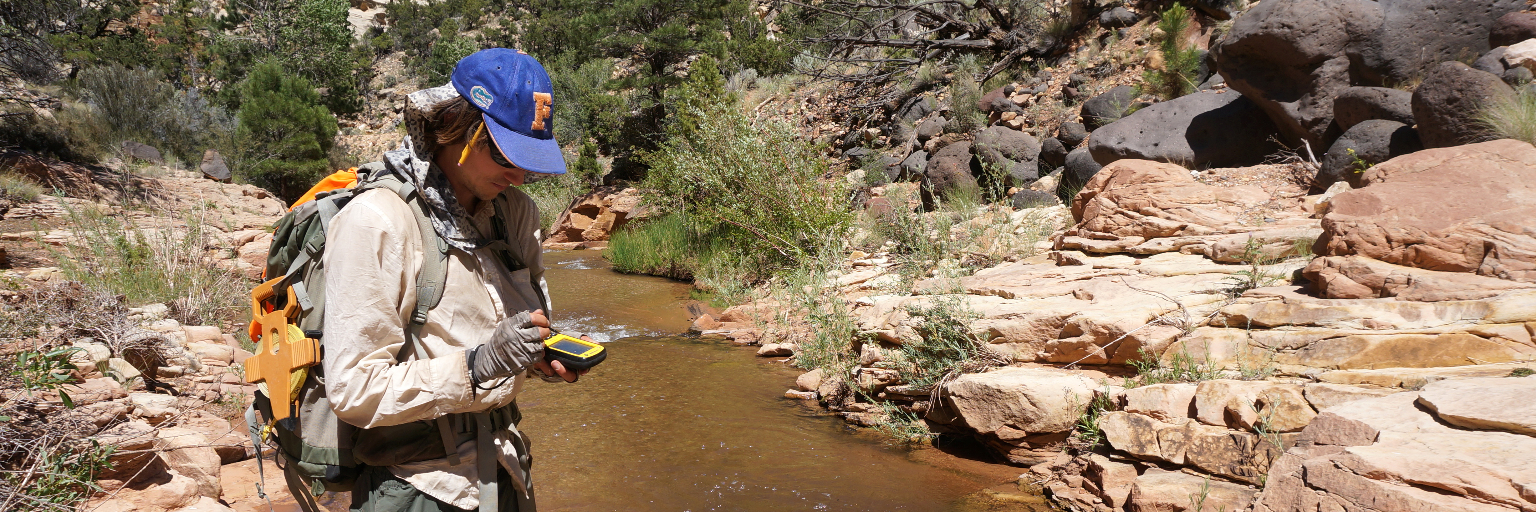 Man wearing backpack with transect tape stands next to a red rock stream using a GPS unit.