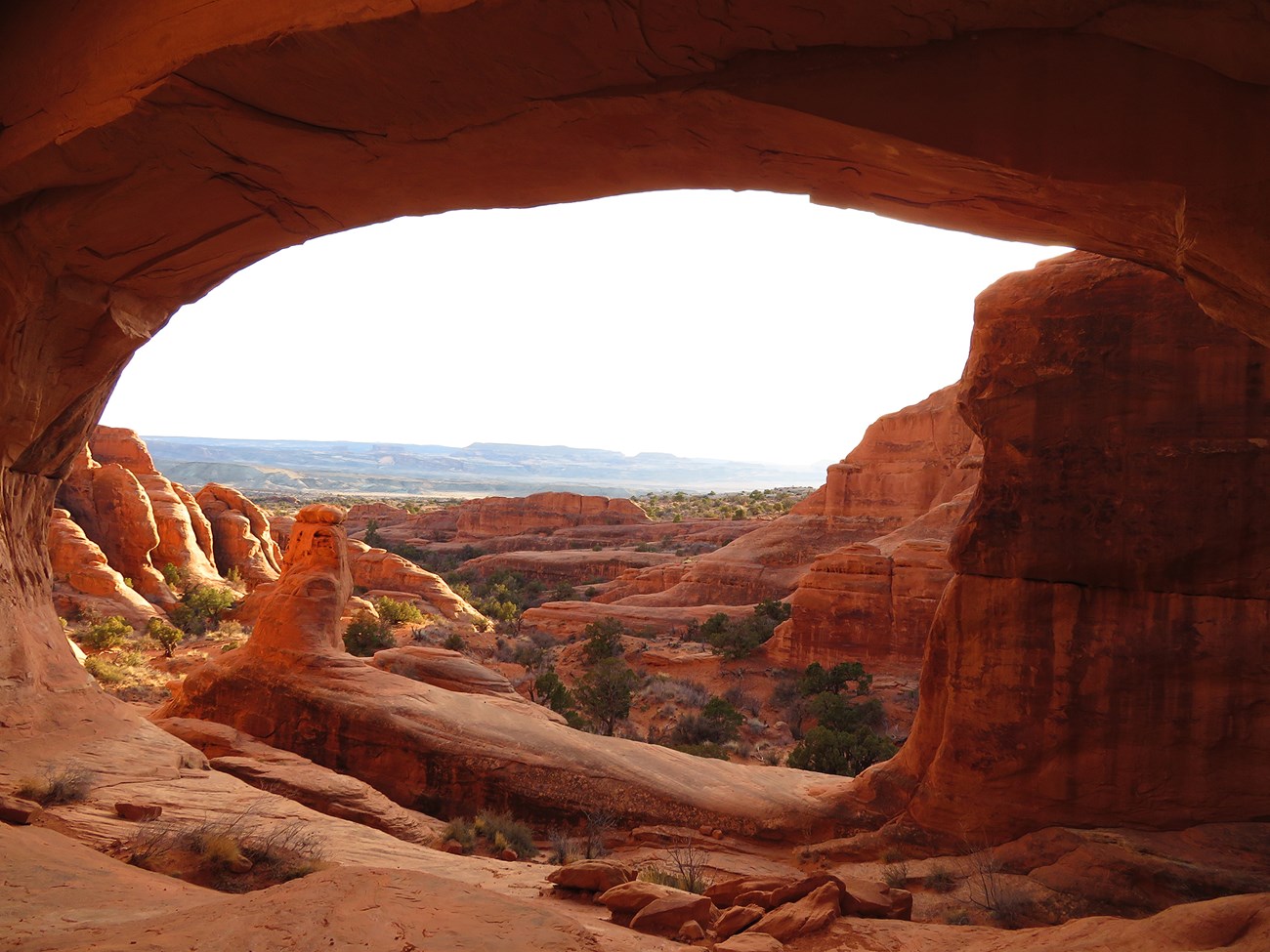 A red sandstone arch overlooks layered rocks, shrubs, and distant mesas under a bright sky.