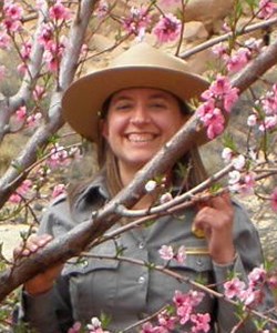 Carolyn Livensperger Woman in NPS uniform stands among pink-flowering fruit trees.