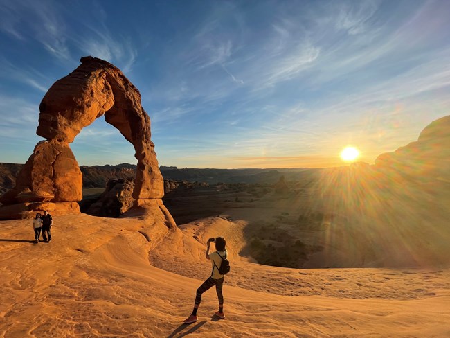 A person taking a photo of a couple with a tall freestanding rock arch in the background and the sun setting.