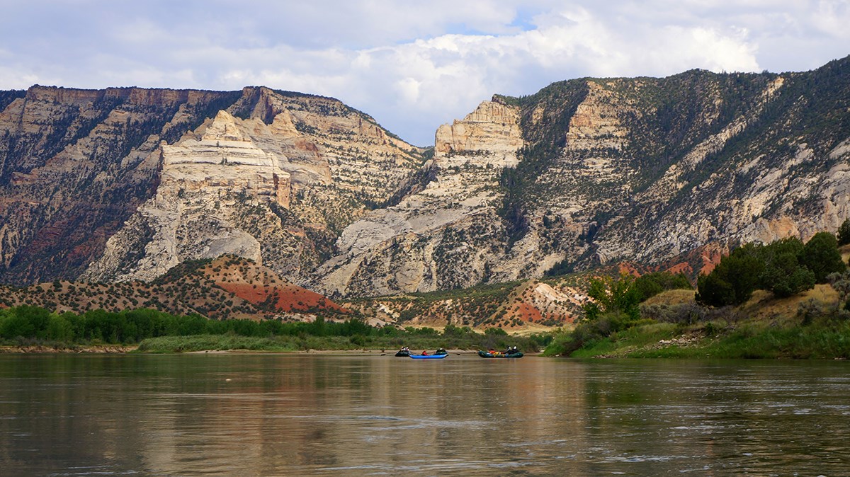 The Green River flows through Dinosaur National Monument, framed by towering rock formations. Cliffs in beige, brown, and red contrast with green riverbanks. A group paddles on calm waters, surrounded by the rugged landscape.