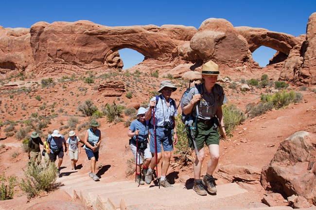 Ranger-led walk at The Windows NPS Andrew Kuhn Ranger and group of hikers ascending trail near sandstone arches at Arches National Park.