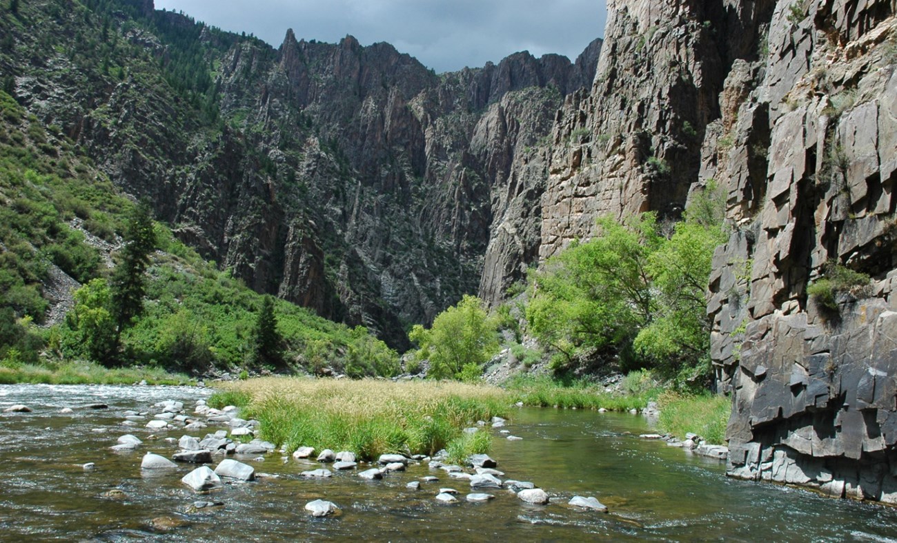 Patch of grass grows on island in river, towering grey cliffs overhead.