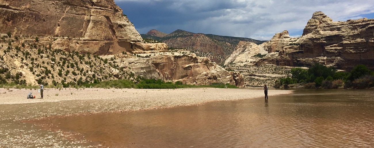 Two people stand in and near a clear river flowing over brown rock and sand.