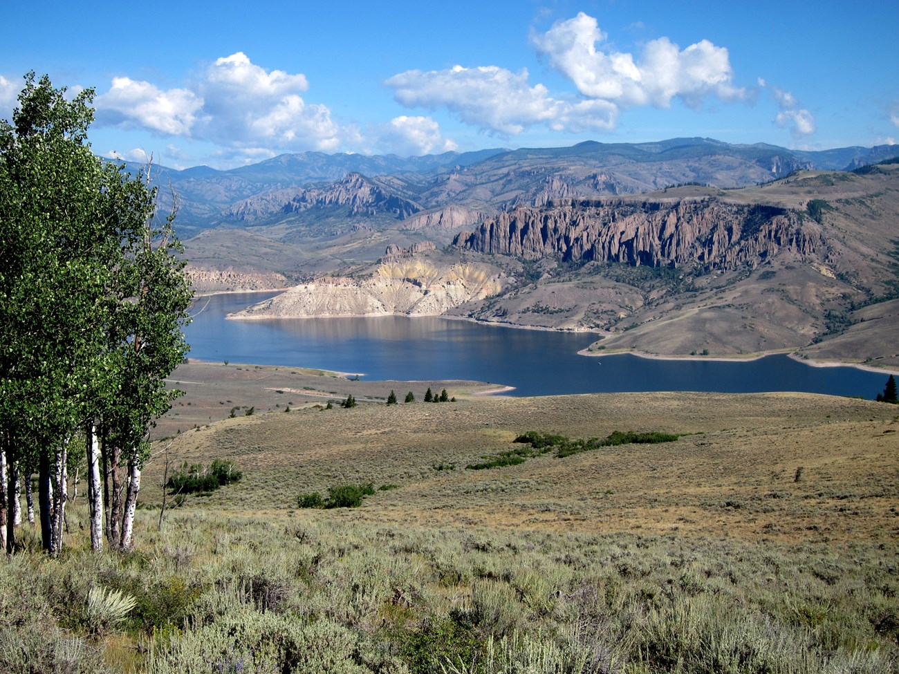 Blue Mesa Reservoir stretches beneath rugged cliffs and rolling sagebrush at Curecanti National Recreation Area. View of Blue Mesa Reservoir surrounded by cliffs, sagebrush, and aspens under a blue sky.